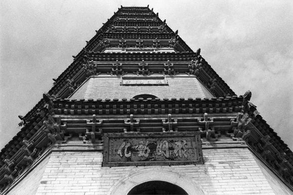 Ilford Pan 400 (FF)

English
A black-and-white close-up photograph of the upper section of a traditional Chinese pagoda. The image focuses on the intricate wooden eaves and decorative elements. There is a plaque with Chinese characters above an arched doorway. The sky is bright in the background.
中文
一张黑白特写照片，拍摄的是中国传统宝塔的上部结构。图片聚焦于精美的木质屋檐和装饰元素。在拱形门洞上方有一块刻有中文字的匾额。背景是明亮的天空。