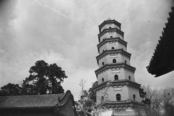 Ilford Pan 400 (FF)

English
A black-and-white photograph of a tall, multi-tiered traditional Chinese pagoda. The pagoda has several levels, each with arched windows and intricate eaves. The structure is framed by trees and a partially visible tiled roof in the foreground. The sky above is mostly clear with some wispy clouds.
中文
一张黑白照片，拍摄的是一座高大的多层中国传统宝塔。宝塔有多个层级，每层都有拱形窗户和精美的屋檐。建筑被树木和前景中部分可见的瓦屋顶所环绕。上方的天空大部分晴朗，点缀着一些缕缕白云。