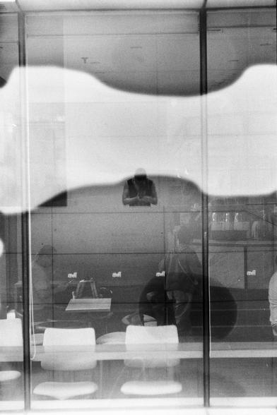 ERA 100 (FF)

English
A black-and-white photograph showing the interior of a modern café or restaurant. The image is taken through a glass window, capturing reflections and distortions. Inside, several people are seated at tables, engaged in conversation or eating. The furniture includes light-colored chairs and tables. The background features large windows and a tiled wall.
中文
一张黑白照片，展示了一家现代咖啡馆或餐厅的内部。照片通过玻璃窗拍摄，捕捉到反光和扭曲的影像。室内有几个人坐在桌旁，正在交谈或用餐。家具包括浅色的椅子和桌子。背景是大窗户和瓷砖墙。