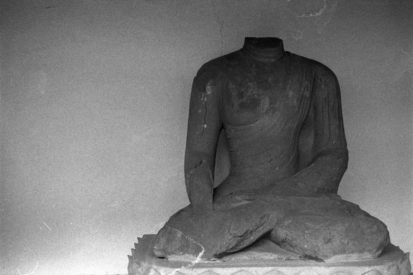 Ilford Pan 400 (FF)

English
A black-and-white photograph of a headless stone statue of a seated figure. The statue is in a meditative pose, with legs crossed and hands resting on the lap. The figure is draped in a robe that covers both shoulders and falls to the knees. The statue is positioned against a plain, slightly textured wall. The surface of the statue shows signs of wear and damage, particularly around the neck and torso.
中文
一张黑白照片，拍摄的是一尊无头的坐姿石像。石像呈冥想姿势，双腿交叉，双手置于腿上。人物身披长袍，覆盖双肩并垂至膝部。石像背…