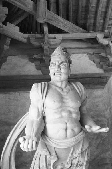 Ilford Pan 400 (FF)

English
A black-and-white photograph of a stone statue of a muscular, standing figure. The figure has a fierce expression, with a wide-open mouth and prominent eyes. It is dressed in a detailed robe with intricate patterns near the waist. The statue is positioned under a wooden structure with visible beams and brackets. The figure's right arm is raised with a clenched fist, while the left arm is bent at the elbow, hand resting on the hip.
中文
一张黑白照片，拍摄的是一尊肌肉发达的站立石像。人物面部表情凶猛，…