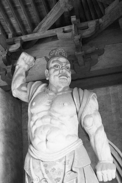 Ilford Pan 400 (FF)

Ilford Pan 400 (FF)

English
A black-and-white photograph of a stone statue of a muscular, standing figure. The figure has a fierce expression, with a wide-open mouth and prominent eyes. It is dressed in a detailed robe with intricate patterns near the waist. The statue is positioned under a wooden structure with visible beams and brackets. The figure's left hand is extended forward, palm facing down, clenched, while the right hand is raised with a clenched fist.
中文
一张黑白照片，…