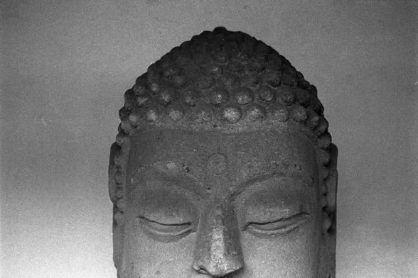 Ilford Pan 400 (FF)

English
A black-and-white close-up photograph of the head of a stone Buddha statue. The statue features a serene expression with closed eyes and a slightly smiling mouth. The head is adorned with tightly curled hair, arranged in a pattern of small, rounded bumps. The surface of the statue appears smooth and well-preserved.
中文
一张黑白照片，特写拍摄的是一尊石佛头像。佛像面部表情宁静，双目微闭，嘴角略带微笑。头部装饰有卷曲的发髻，呈小而圆的凸起状排列。石像表面看起来光滑且保存完好。