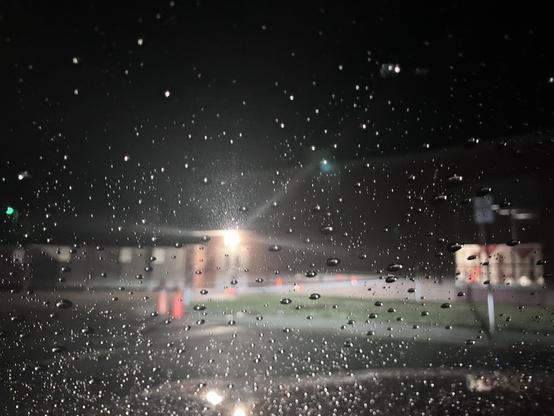 A close-up view of raindrops on a car windshield at night, with blurred street lights and traffic cones visible in the background.