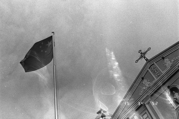 Ilford Pan 400 (FF)

English
Black-and-white photograph of a flagpole with a dark flag featuring a star pattern, flying against a cloudy sky. The top of the church facade, including a cross, is visible on the right side of the image. The timestamp "15 34 52" is visible in the bottom right corner.
中文
黑白照片，旗杆上悬挂着一面带有星形图案的深色旗帜，背景是多云的天空。教堂正面的顶部，包括一个十字架，在图像右侧可见。右下角可见时间戳“15 34 52”。