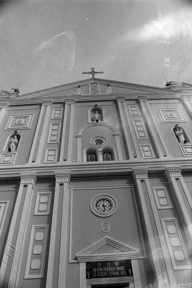 Ilford Pan 400 (FF)

English
Black-and-white photograph of the church facade, taken from a low angle. The building features a pediment with a cross at the top, columns, and statues in niches. A circular clock is visible below the pediment, with the inscription "DOM IN HON DEIPARAE IMMAC SS JOSEPH EFRAN ASS" beneath it. The timestamp "28" is visible in the top right corner.
中文
黑白照片，从低角度拍摄的教堂正面。建筑物顶部有一个山形墙，上面有一个十字架，还有柱子和壁龛中的雕像。山形墙下方可见一个圆形时钟，其下方有拉丁文铭文“DOM IN HON DEIPARAE IMMAC SS JOSEPH EFRAN ASS”…