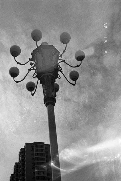 Ilford Pan 400 (FF)

English
Black-and-white photograph of a multi-lamp streetlight. The lamp post is tall and ornate, with seven spherical lamps extending from curved arms. The background features a cloudy sky and the top of a multi-story building, partially obscured by the streetlight.
中文
黑白照片，展示了一盏多灯街灯。灯柱高大且装饰精美，七个球形灯泡从弯曲的灯臂上延伸出来。背景是多云的天空和一栋多层建筑的顶部，部分被街灯遮挡。
