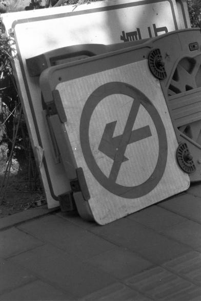 Ilford FP4 Plus 125 (FF)

English
A black-and-white photograph of a tilted, rectangular traffic sign lying on a paved surface. The sign features a circular symbol with a diagonal line crossing through a left-turning arrow, indicating "No Left Turn." The sign is partially obscured by shadows from nearby objects, including what appears to be a plastic chair and other indistinct items.
中文
一张黑白照片，显示一个倾斜的矩形交通标志牌平躺在铺设的地面上。标志牌上有一个圆形符号，一条对角线穿过一个向左转的箭头，表示“禁止左转”。标志牌部分被附近物体的阴影遮挡，包括一个看起来像塑料椅子的物体和其他模糊的物品。