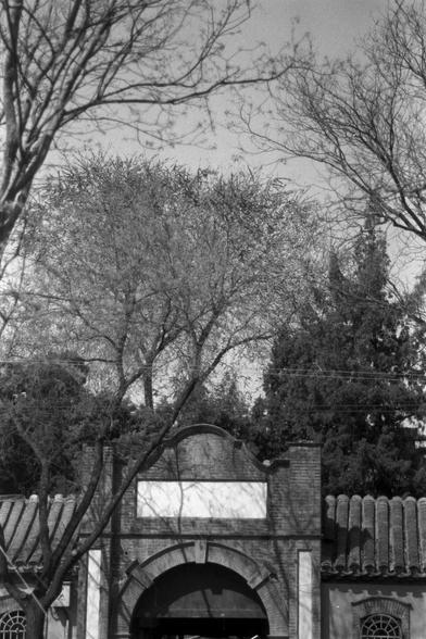 Ilford FP4 Plus 125 (FF)

English
A black-and-white photograph of a traditional arched gateway with a tiled roof. The gateway is made of brick and stone, featuring a rectangular blank space above the arch. Bare trees with thin branches frame the top of the image, and a tall evergreen tree is visible in the background.
中文
一张黑白照片，展示一个传统的拱形门楼，屋顶铺有瓦片。门楼由砖石建造，拱门上方有一个矩形的空白区域。光秃秃的树枝在图片上方形成框架，背景中可见一棵高大的常绿树。