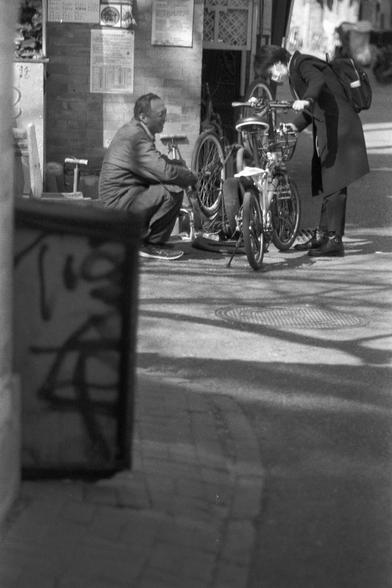 Ilford FP4 Plus 125 (FF)

English
A black-and-white photograph showing a street scene. An older man in a suit sits, facing sideways from the camera, with his hands resting on his knees. Several bicycles are parked, and another person in a coat stands near the bicycles, seemingly engaged in conversation. The background features a brick wall with posters and a manhole cover on the pavement. Shadows of trees and buildings are visible on the ground.
中文
一张黑白照片，展示了一条街道的场景。一位身穿套装的老年男子坐着，侧对镜头，双手放在膝盖上。另…