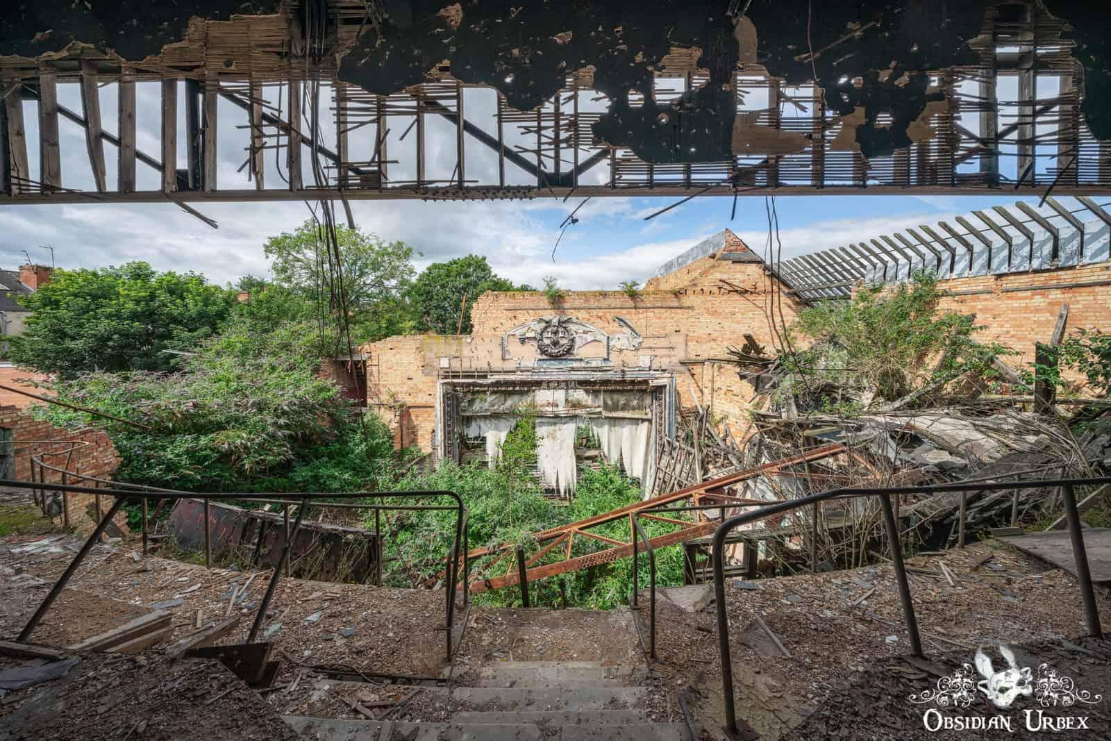 A wide view from an upper level of a severely decayed, overgrown abandoned theatre