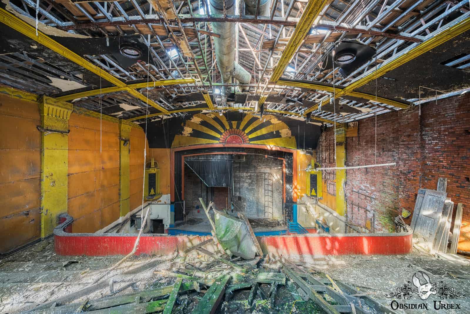 An abandoned Art Deco cinema interior, showcasing a collapsed ceiling, debris, and a vibrant sunburst stage design.