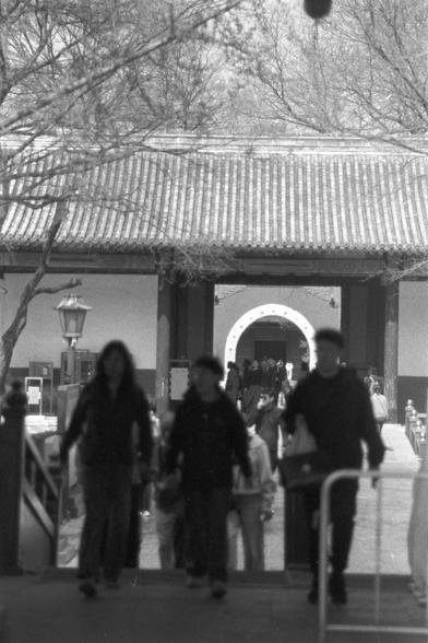 Ilford FP4 Plus 125 (FF)

##English
Black-and-white photograph of a traditional Chinese architectural entrance. The scene features a tiled roof with intricate eaves, supported by wooden pillars. A large, circular doorway is visible in the background, framed by a white arch. Several people, some blurred due to motion, walk in front of the entrance. Bare tree branches are visible above, and a classic streetlamp stands to the left.
##中文
这张黑白照片展示了一个传统中式建筑的入口。画面中有精美的屋顶瓦片和木制柱子支撑的屋檐。背景中有一个巨大的圆形门洞，被白色拱…