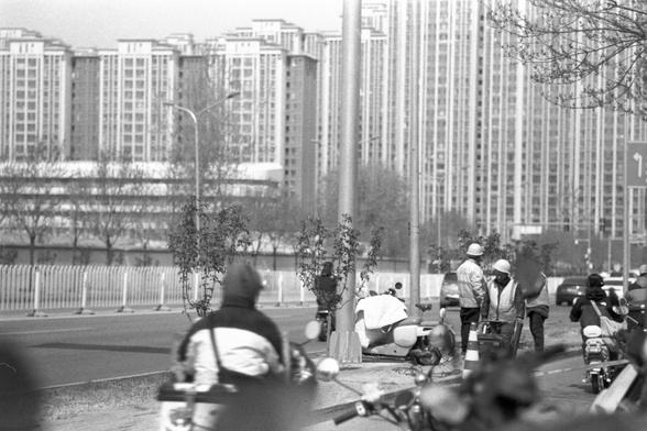 Ilford FP4 Plus 125 (FF)

##English
Black-and-white photograph of a modern urban street scene. In the foreground, several motorcyclists and pedestrians are visible, some wearing helmets and reflective vests. A row of young trees lines the sidewalk, and a tall fence runs parallel to the road. In the background, a cluster of high-rise apartment buildings with vertical lines and balconies dominates the skyline.
##中文
这张黑白照片展示了现代城市街道的场景。前景中有几位摩托车骑手和行人，部分人戴着头盔和反光背心。人行道上种植了一排年轻的树木，一道高高的围栏与道路平行。背景中，一群高…