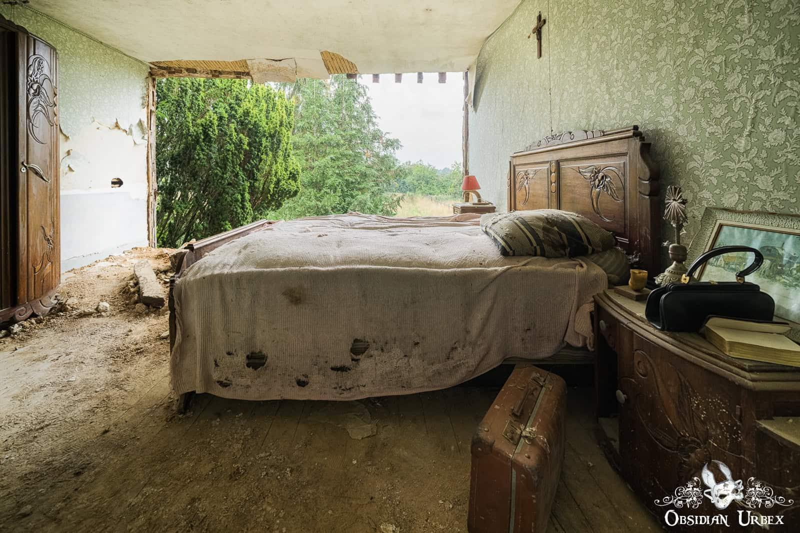An abandoned bedroom in a French farmhouse features a bed, nightstand, and suitcase, looking out through a collapsed wall to trees.