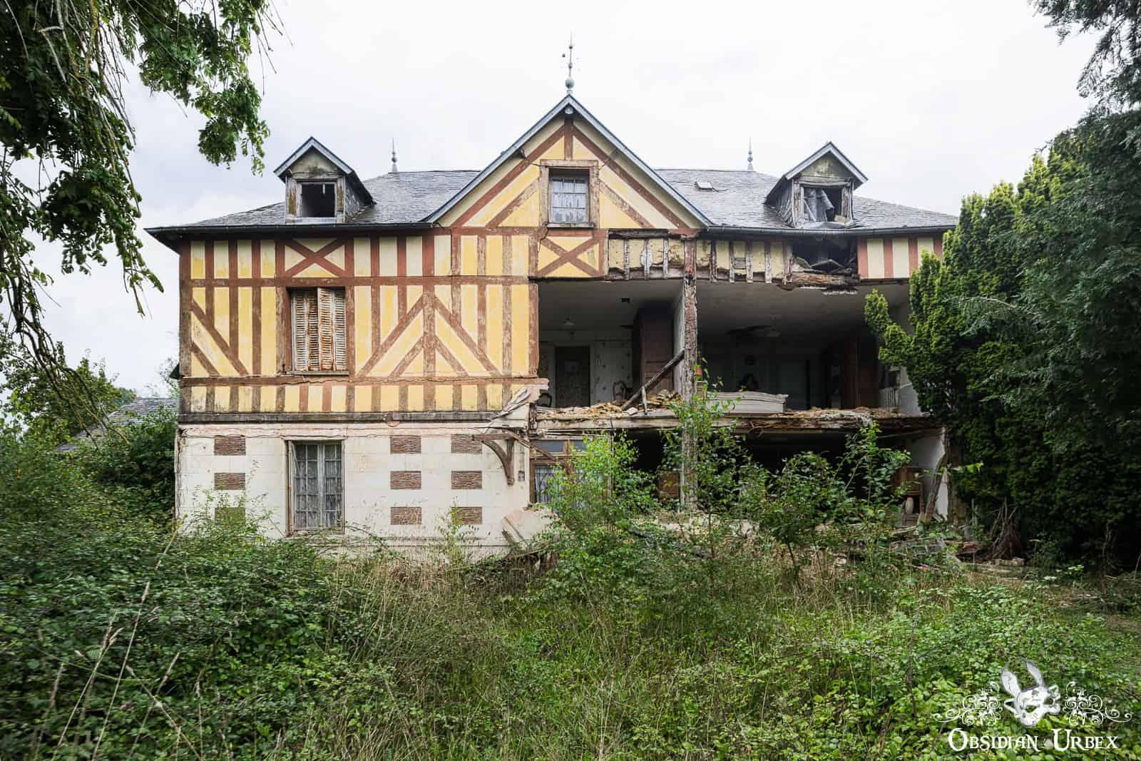 An abandoned, half-timbered farmhouse in France is collapsing, with a large section of its facade missing.