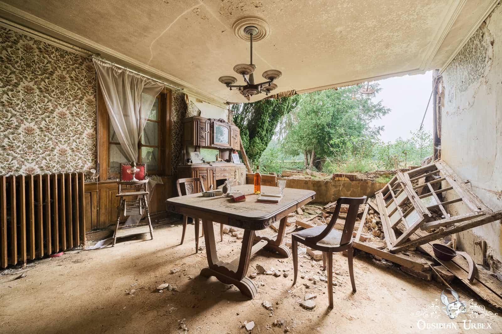 An abandoned French dining room with a collapsed wall opening to nature.