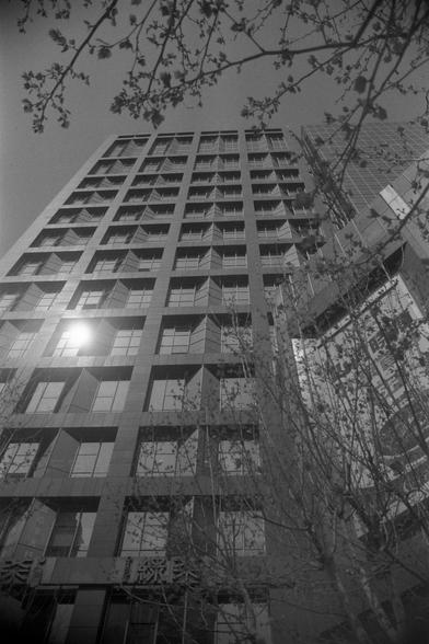 Ilford FP4 Plus 125 (FF)

English
A black-and-white photograph of a tall, modern high-rise building with numerous windows. The perspective is from a low angle, looking up towards the sky, with bare tree branches framing the top of the building. The sun is visible near the top, creating a bright reflection on the building's facade.
中文
一张黑白照片，拍摄的是一幢高大的现代高层建筑，有很多窗户。视角从低角度向上拍摄，望向天空，光秃秃的树枝框住了建筑的顶部。太阳在顶部附近可见，在建筑的立面上形成明亮的反光。