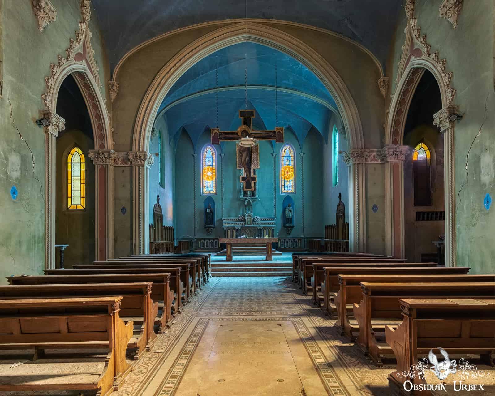 An atmospheric view inside an abandoned blue Italian chapel. Wooden pews lead to an altar with a large suspended wooden cross.