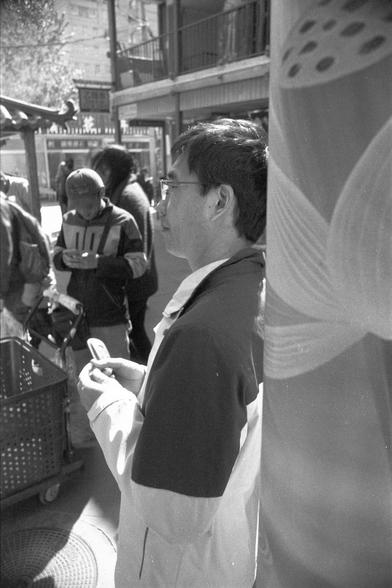 Ilford FP4 Plus 125 (FF)

English
A black-and-white street scene featuring a man in the foreground, wearing glasses and a jacket. He stands near a large cylindrical pillar with a decorative pattern. In the background, other people are visible, some holding shopping baskets, and buildings with signage line the street.
中文
一张黑白的街道场景照片，前景是一名戴眼镜、穿夹克的男子。他站在一个带装饰图案的大圆柱旁。背景中可以看到其他人，有些人拿着购物篮，街道两侧是带有招牌的建筑。