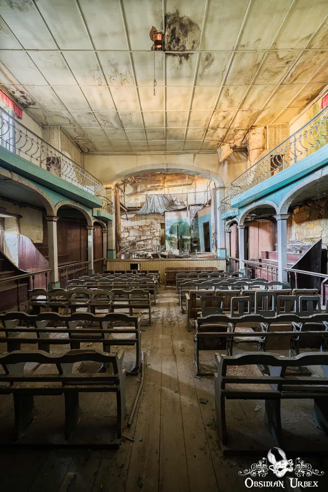 An abandoned theater features rows of worn wooden seats facing a decaying stage, with elevated balconies flanking both sides. The room shows signs of extensive water damage and neglect.