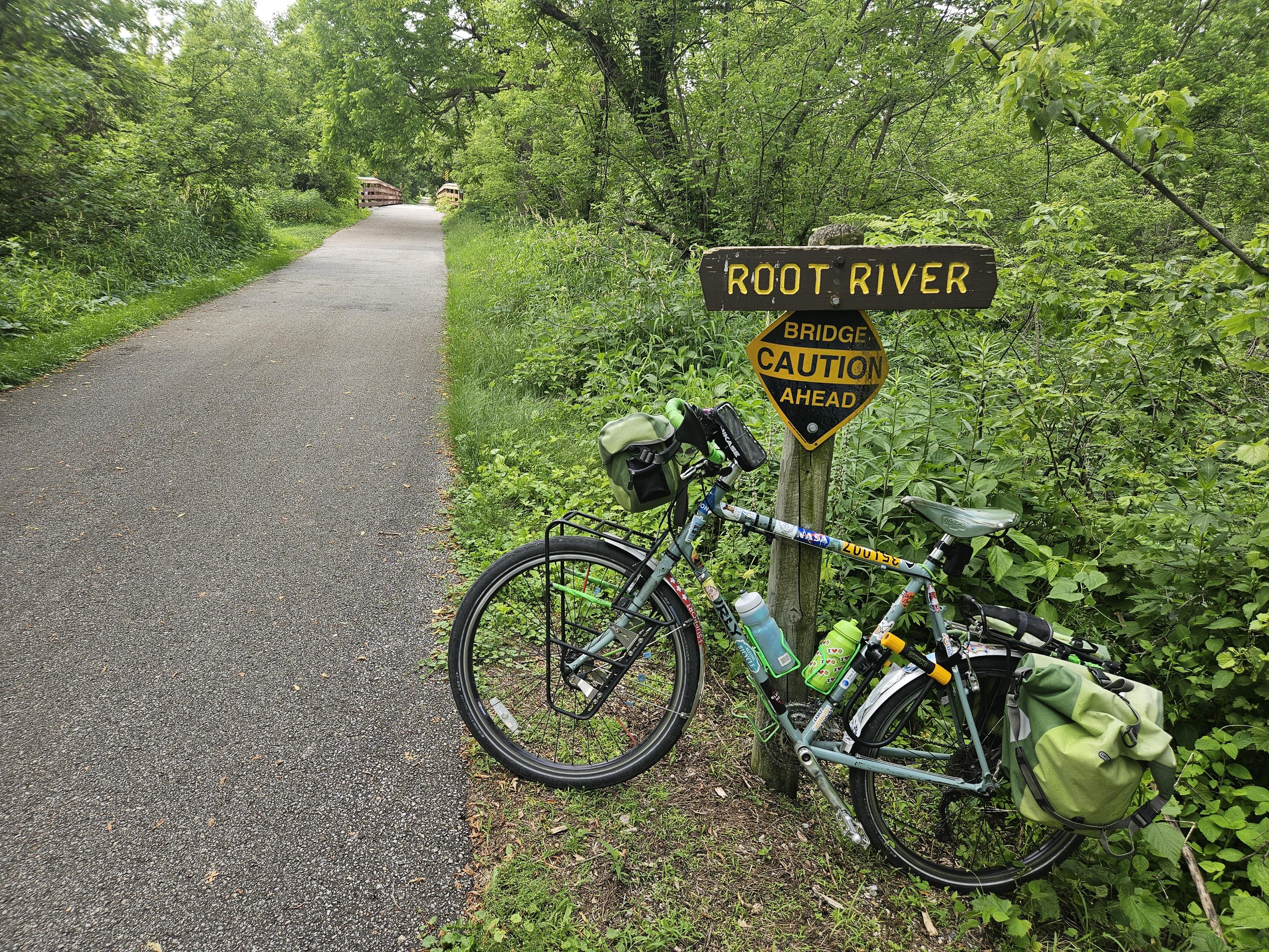 A bicycle with green bags leans against a sign that says Root River bridge caution ahead. It is next to an asphalt path surrounded by green grass,  bushes and trees with green leaves. Brown wood railing fir a bridge can be seen in the distance.