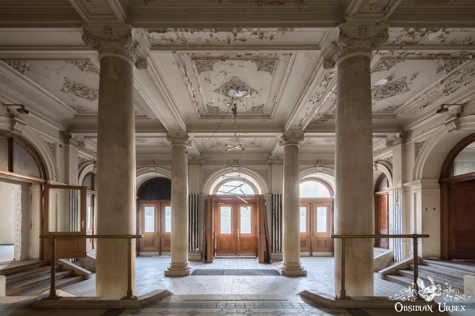An ornate, decaying entrance foyer with tall columns, peeling ceilings, and wooden doors.