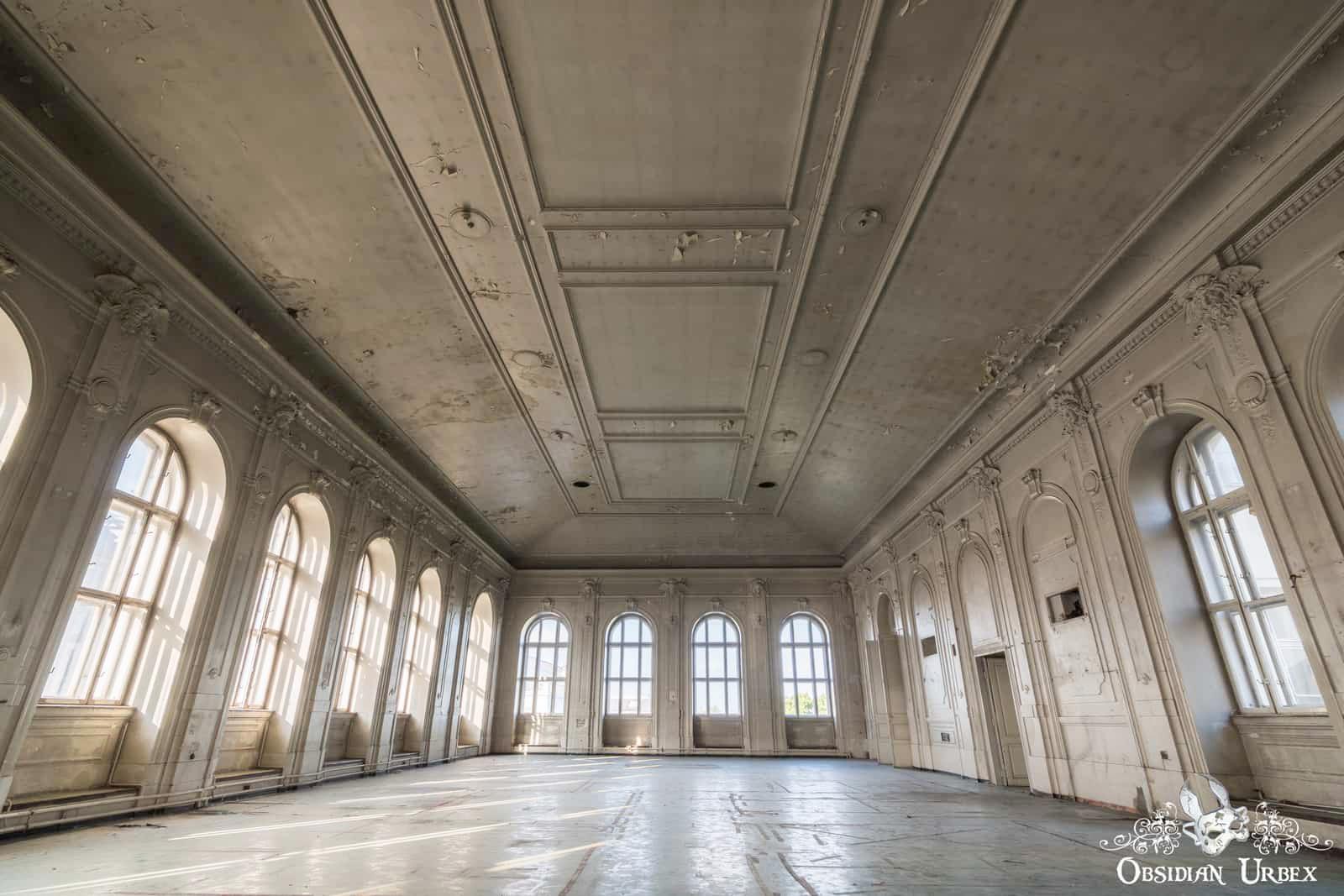 A grand, abandoned hall features high, ornate ceilings with peeling paint, and numerous arched windows illuminating its dusty floor