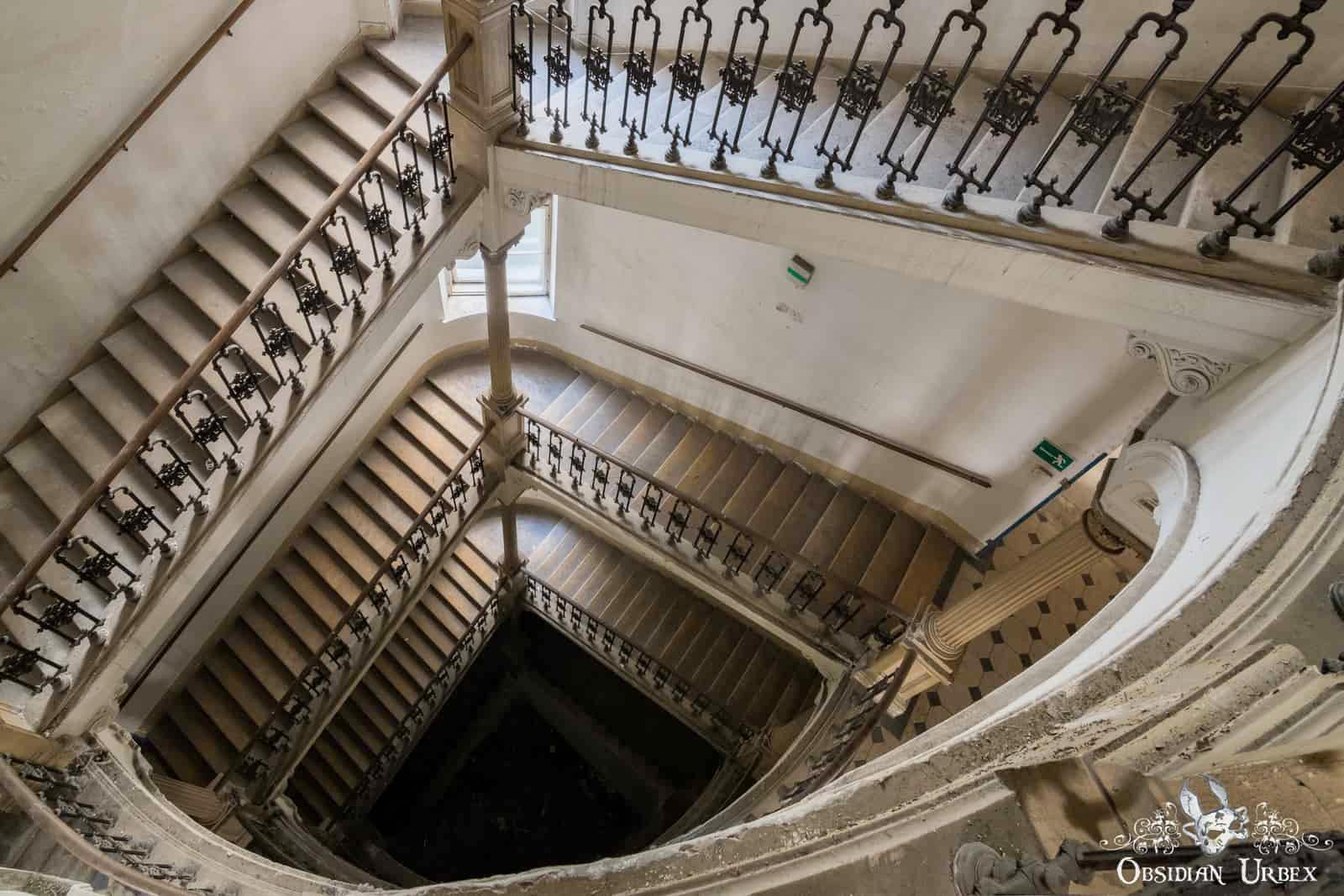 Looking down into a vast, abandoned staircase with ornate iron railings and wooden steps spiraling several floors into darkness.