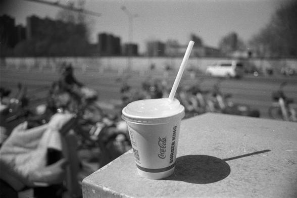 Ilford FP4 Plus 125 (FF)

English
A black-and-white photo featuring a Burger King paper cup with a lid and a straw placed on a concrete ledge. The cup is branded with the Burger King logo and text. In the background, there is a blurred urban scene with buildings, trees, and a crowd of people, suggesting a public outdoor setting.
中文
一张黑白照片，画面中有一个放在混凝土台面上的汉堡王纸杯，杯子带盖和吸管。杯身印有汉堡王的标志和文字。背景是模糊的城市景观，有建筑、树木和人群，暗示这是一个户外公共场所。