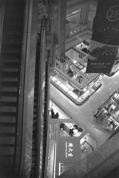 Ilford FP4 Plus 125 (FF)

English
A black-and-white photo taken from an elevated position inside a shopping mall. The image shows escalators, glass railings, and multiple floors with various stores. Signs in Chinese characters, including "Chow Tai Seng" and "LAOMIAO," are visible. The mall is well-lit and features a modern architectural design.
中文
一张从购物中心高处拍摄的黑白照片。画面中有自动扶梯、玻璃栏杆和多个楼层的各类店铺。可以看到中文字样的标识，包括“周大生”和“老庙”。商场灯光明亮，建筑设计现代。