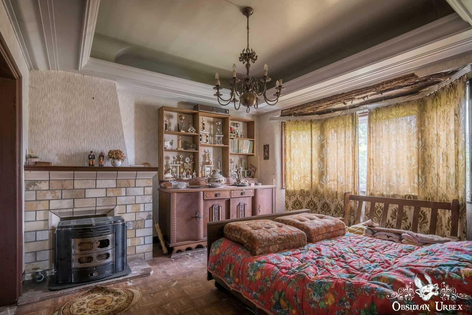 An abandoned bedroom-like living room shows a prominent bed, fireplace, and a grand bookcase full of pigeon racing trophies.