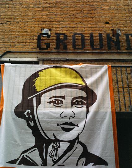 Harman Phoenix 200 II (6x7)

##English
A large banner hangs on a brick building, displaying a stylized colour illustration of a worker wearing a helmet. Above the banner, the word "GROUND" is partially visible in bold, black letters.
##中文
一栋砖墙建筑上悬挂着一幅大型横幅，上面是一位戴安全帽的工人人的画像。横幅上方，“GROUND”几个字母部分可见，字体粗体黑色。