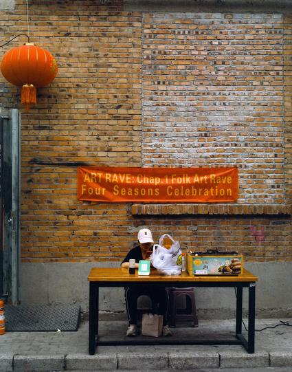 Harman Phoenix 200 II (6x7)

##English
A person sits at a wooden table under an orange banner that reads: "ART RAVE: Chap. 1 Folk Art Rave Four Seasons Celebration." The table holds a plastic bag, a drink, a box of food, and a small stool. A red lantern hangs on the left side of a brick wall in the background.
##中文
一人坐在木桌前，桌上方悬挂着一条橙色横幅，上面写着：“ART RAVE: Chap. 1 Folk Art Rave Four Seasons Celebration”。桌上放着一个塑料袋、一杯饮料、一个食物盒和一个小凳子。背景是砖墙，左侧挂着一个红灯笼。