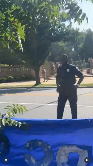 Video shot from the protest crowd outside Malcolm X Park. In the foreground, you see a blue protest banner being held by the people. In white letters in this part it says "DC." A DC police officer is seen in front of the crowd, but just standing there, doesn't look like an intimidation attempt. In the background you see National Guard troops-- who should be home with their families!-- standing around the perimeter of the park.