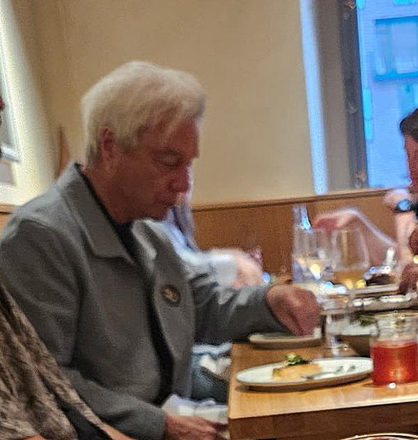 An older man with gray hair that flops across his head from left to right, wearing a gray jacket, looking down at a plate of food with one hand on his lap and the other reaching for something near/on the plate. He's sitting at a dining table on which sits a glass with red liquid in it and what appears to be a stemmed glass of white wine. There is another table beyond it. I'm no expert, but it appears he's having the grilled waffle with smoked salmon, trout roe and yogurt.