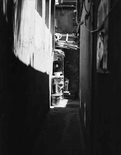 Lucky LUCKY SHD 400 (6x7)

English
A black-and-white photograph of a narrow alleyway. The alley is dimly lit, with sunlight streaming in from the right, casting sharp shadows on the walls and ground. Various objects, including buckets, a ladder, and other indistinct items, are stacked or leaning against the walls. The alley appears cluttered and leads into darkness at the far end.
中文
一张黑白照片，展现了一条狭窄的小巷。小巷光线昏暗，右侧有阳光照射进来，在墙壁和地面上投下清晰的阴影。各种物品，包括水桶、梯子和其他难以辨认的物品，堆放或靠在墙壁上。小巷看起来杂乱无章，远端延伸至黑暗中。