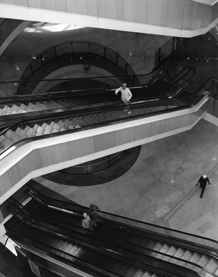Lucky LUCKY SHD 400 (6x7)

English
A high-contrast black-and-white photograph of a modern shopping mall interior. The scene is dominated by multiple escalators with smooth, curved handrails and metallic steps. Some individuals are visible: one person stands on the upper-level escalator, another walks near the base of the escalators, and two are midway on a lower escalator. The floor and walls are tiled, and the ceiling features a large, circular skylight allowing natural light to filter in. The…