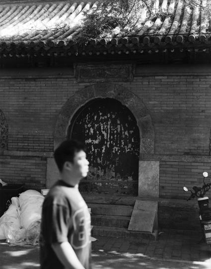 Lucky LUCKY SHD 400 (6x7)

English
A black-and-white photograph capturing a traditional Chinese architectural structure. The building features a tiled roof with intricate eaves and a brick wall with a large, arched doorway. The doorway is sealed with wooden boards, and there is a stone plaque with Chinese characters above it. A person in a light-colored shirt is walking in the foreground, slightly blurred. The ground is paved with bricks, and there are bags or sacks stacked to the left of the d…