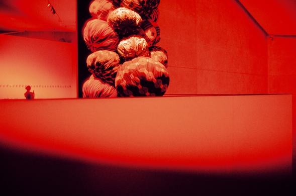 Harman Red 125 (FF)

English
An art installation in a dimly lit, red-toned room. A large, vertical sculpture made of stacked, fabric-covered objects resembling oversized cabbages dominates the space. The walls and floor are bathed in a deep red light, creating a dramatic and immersive atmosphere. A small, blurred figure stands near the sculpture, adding a sense of scale.
中文
一个光线昏暗、红色调的房间内的艺术装置。一个巨大的垂直雕塑由堆叠的、布料覆盖的物体组成，形似放大的卷心菜，主导了整个空间。墙壁和地板浸润在深红色的灯光中，营造出戏剧性和沉浸式的氛围。一个小而模糊的人影站在雕塑附近，增添了尺寸感。