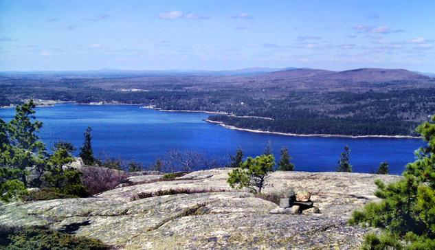 A beautiful sunny early spring day. The sky is a deep robins egg blue with a sprinkling of small clouds, the ocean is a deep blue. Atop a naked gray rock crest with a few scrub pines and smaller leafless vegetation. Hundreds of feet below an arm of the ocean runs inland from our right to left. Across this sound is the other half of this island, featuring several impressive mountains and a few vacation homes along the far shore. The majority of the visible land below is wooded, mainly pines and other evergreens. The hardwoods are still leafless and the grass is just starting to turn green.