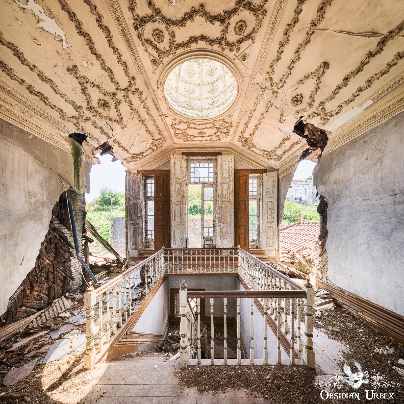 An abandoned grand hall featuring a collapsing wooden staircase and an ornate, damaged ceiling with a skylight. Crumbling walls reveal views outside.