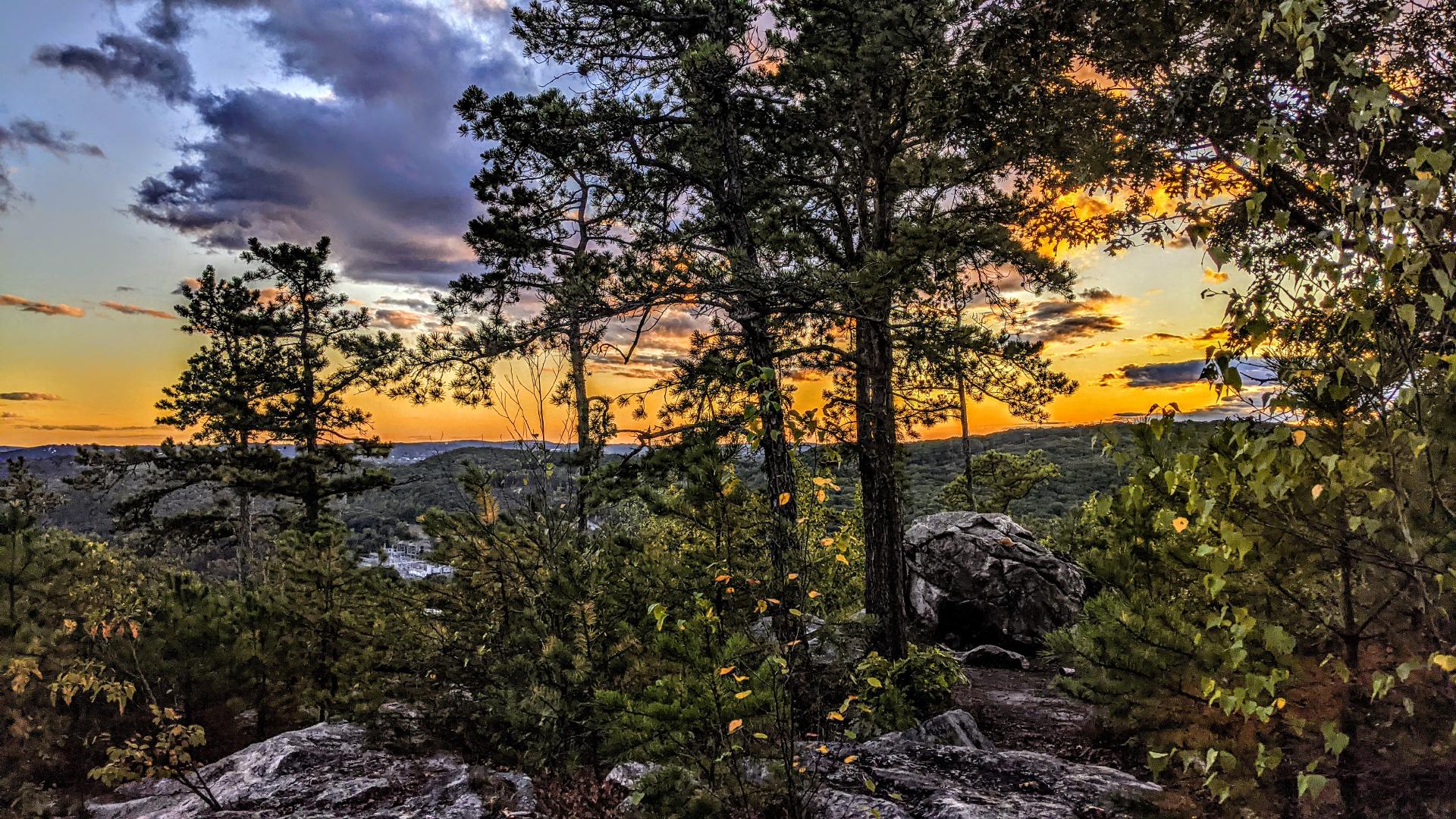 A pale blue gray sky with dark purple gray clouds drifting past. The horizon and the edges of nearby clouds are tinted gold, orange and yellow by the setting sun. We are standing atop a cliff top of light gray rock streaked with darker gray lichen. The rocky ground is sprinkled with weather-beaten pines and leafy shrubs just beginning to turn gold for autumn. A few hundred feet below are the outskirts of a small city, a series of low, mostly wooded rolling ridges with a factory and a few other buildings poking through. A few miles off is a slice of a more built-up portion of the city near the golden horizon.