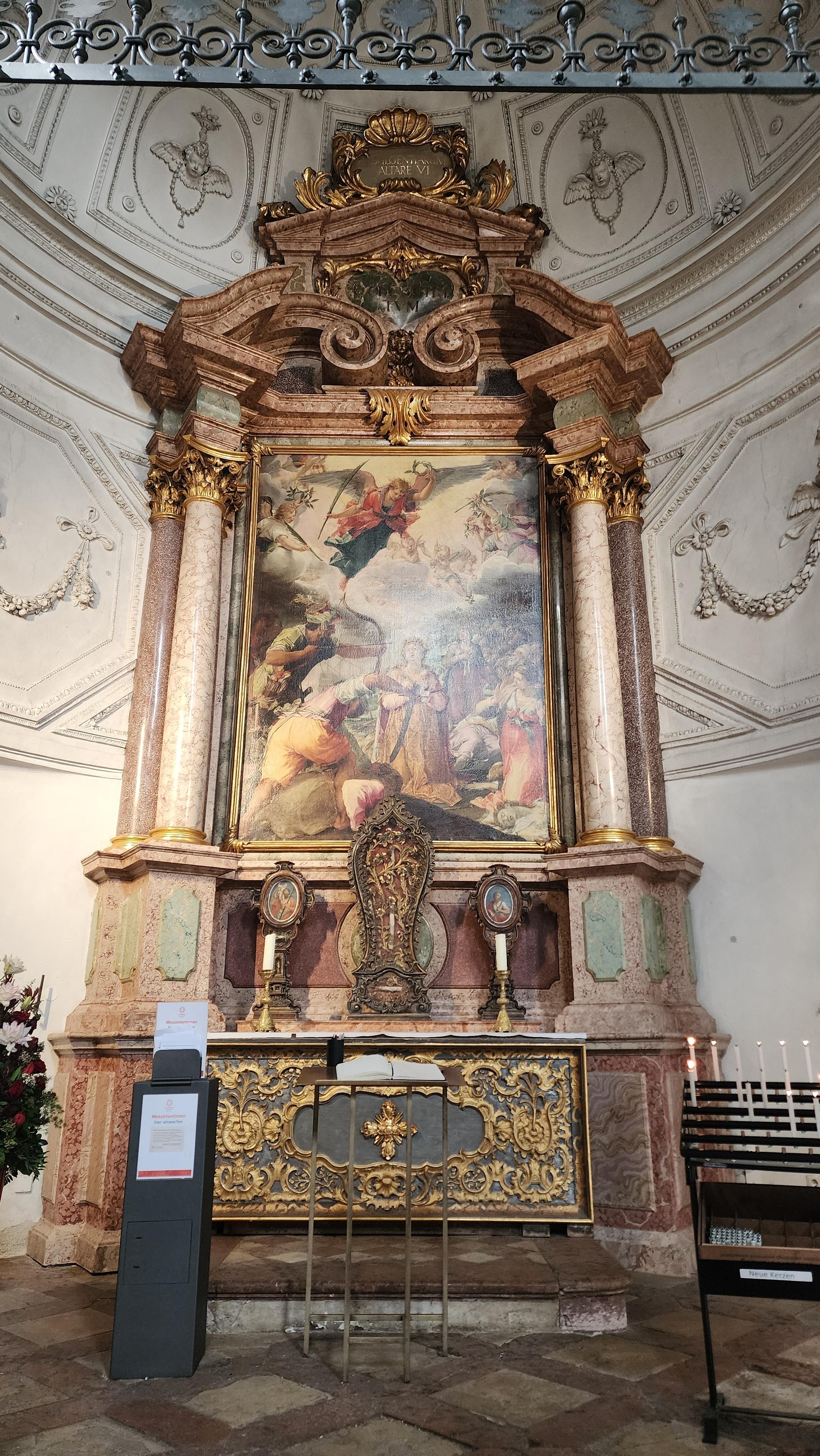Large domed, semicircular side chapel with white walls and an altarpiece about 15-20 ft. tall that appears to be made of red marble, displaying at center a large painting of an angel flying over a group of women who are about to be executed by men brandishing swords and arrows. Below this are three display cases containing bones, the center one taller than the other two. These are sitting above a rectangular altar, on the floor.