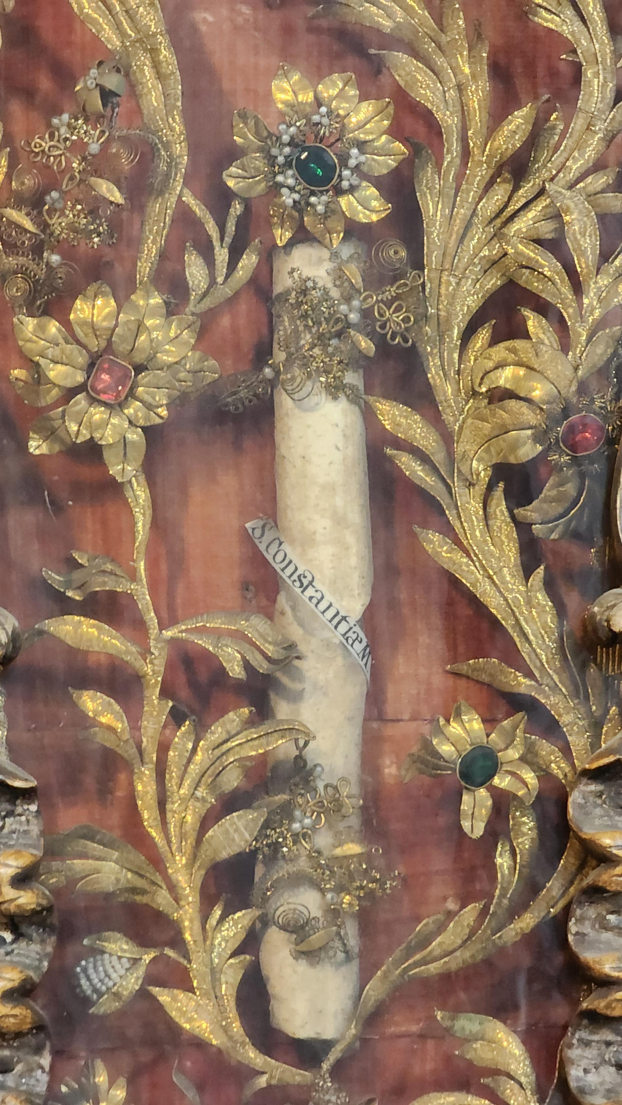 The bone in the central display. A long arm or leg bone with a label around its center reading "S. Constantiae M." The bone is surrounded and held in place by gold and gem floral ornaments.