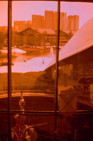 Harman Red 125 (FF)

##English
A sepia-toned view through a window with black frames. Outside, a park with a paved walkway is visible, featuring a person walking by the side of a human shaped statue. In the background, there are tall residential buildings and a large, dark-roofed building surrounded by trees. The foreground inside the window shows part of a metallic object, possibly a lamp or decorative item, with a curved, reflective surface.
##中文
通过黑色框架的窗户，看到一个带有铺砌步道的公园，一个人在一个大型人形雕塑旁行走。远处是高层住…