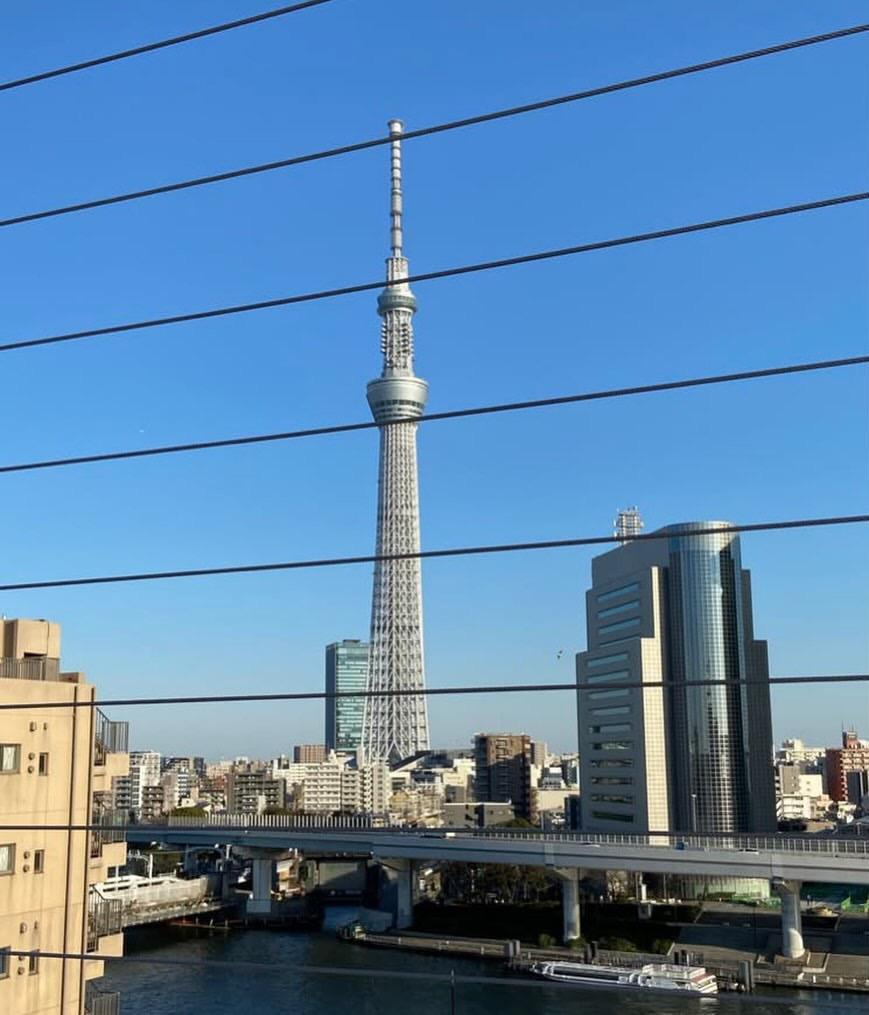 Picture - against a blue sky, the Tokyo Skytree (a very tall TV/radio mast with viewing pods near the top), seen through the window of a train.