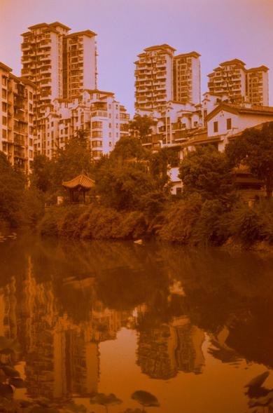 Harman Red 125 (FF)

##English
A sepia-toned image showing a serene urban scene with a pond in the foreground. The pond reflects tall residential buildings and lush green trees. A traditional pavilion with a curved roof is partially visible among the trees.
##中文
一张深褐色调的图像，展示了一个宁静的城市景观，前景是一个池塘。池塘倒映着高耸的住宅楼和茂密的绿树。树丛中部分可见一个传统的亭子，亭顶为弯曲的屋顶。