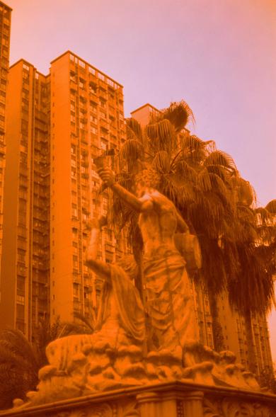 Harman Red 125 (FF)

##English
An orange-tinted photograph featuring a statue of a robed figure holding a torch aloft, surrounded by palm trees. In the background, tall residential buildings with numerous windows and balconies rise against the sky.
##中文
一张橙色调的照片，展示了一尊手持火炬的长袍人物雕像，周围环绕着棕榈树。背景中是高耸的住宅楼，楼上有许多窗户和阳台，矗立在天空下。