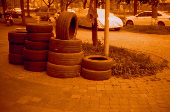 Harman Red 125 (FF)

##English
A reddish-orange tinted image of a sidewalk lined with stacks of used tires. The tires are arranged in multiple stacks, with some lying flat. In the background, there are parked cars, trees, and a covered vehicle.
##中文
一张红橙色调的图像，展示了人行道上堆放的废旧轮胎。轮胎整齐地堆叠成多堆，有些平放着。背景中有停放的汽车、树木和一个被覆盖的车辆。